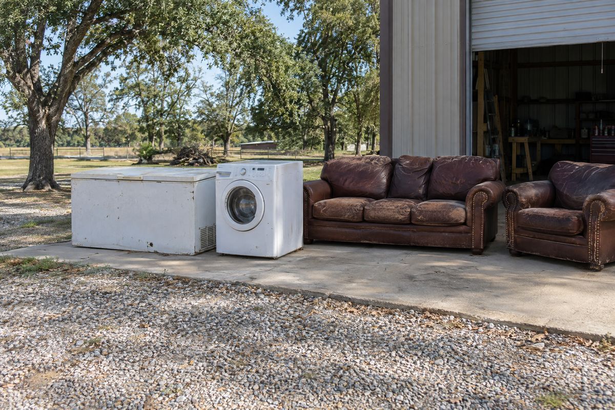 A white chest freezer, a washing machine and a worn leather sofa on the concrete apron in front of a metal shop building