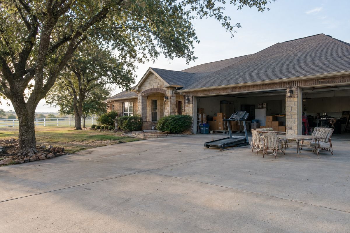 A treadmill, old patio furniture and boxes staged on the wide drive of a stone and brick ranch home, with pecan trees and a horse fence behind