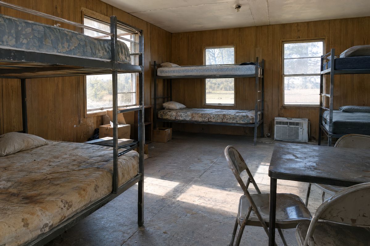 Metal bunk bed frames, stained mattresses, boxes and an old window air conditioner inside a dusty bunkhouse