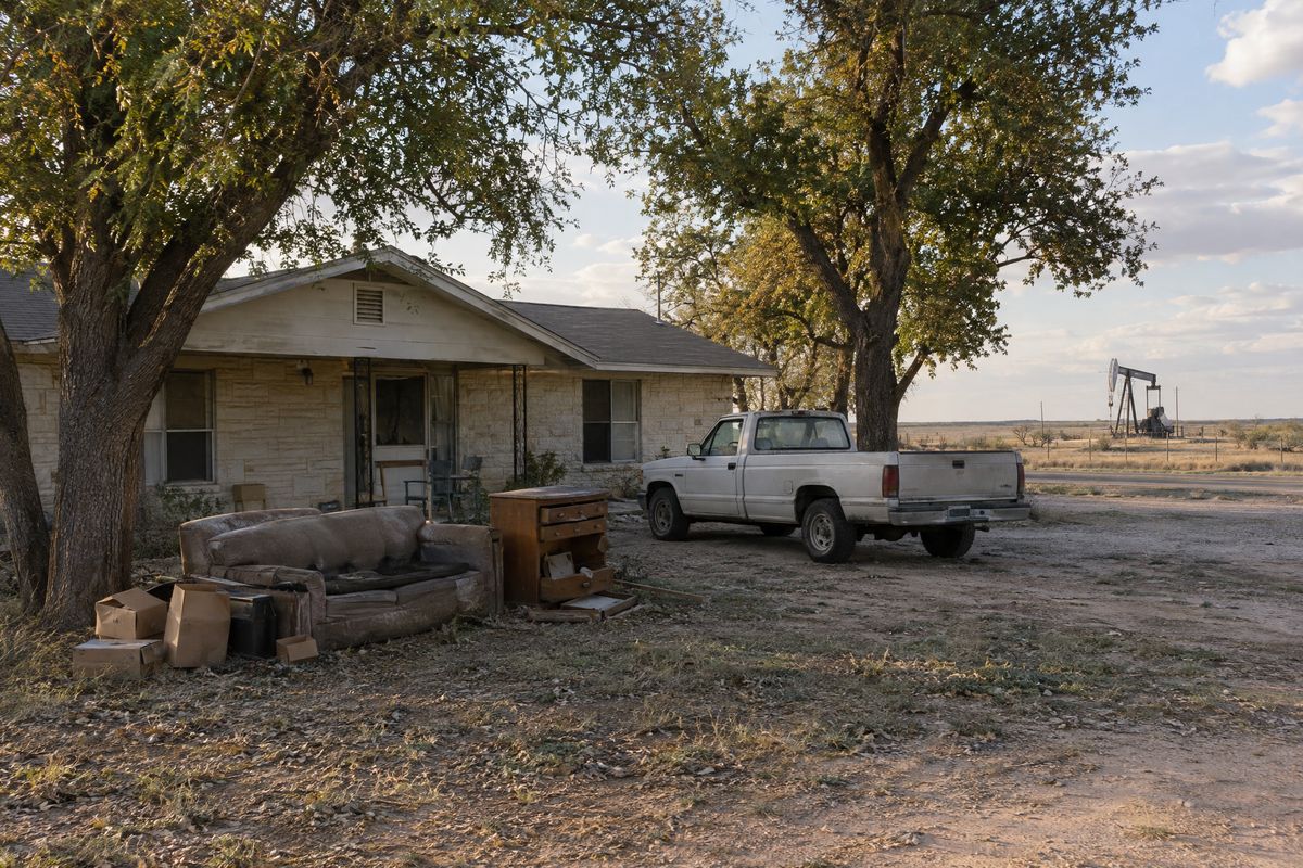 An old couch, boxes and a broken dresser beside a pickup in the yard of a ranch house among pecan trees, with a pumpjack across the road
