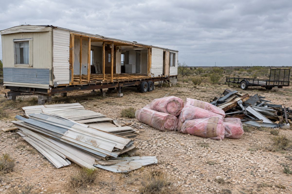 A partly dismantled single-wide mobile home with stacked siding panels, bagged insulation and a separate scrap metal pile on a caliche lot