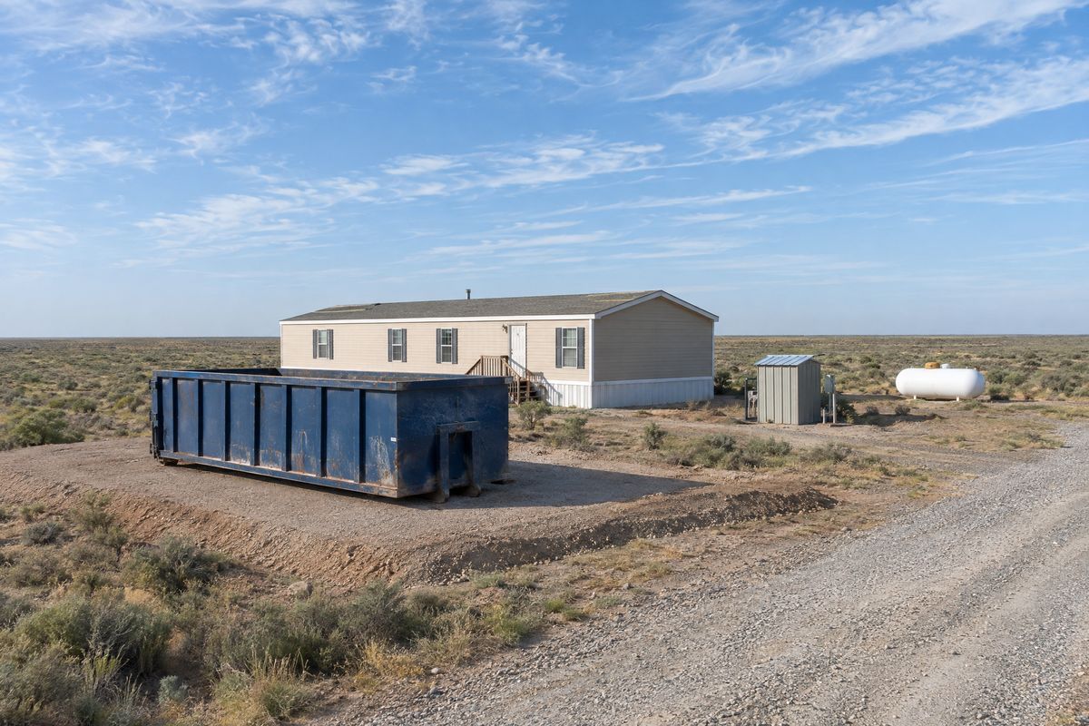 A roll-off container on a caliche pad beside a single-wide manufactured home, with a small well enclosure and a propane tank nearby