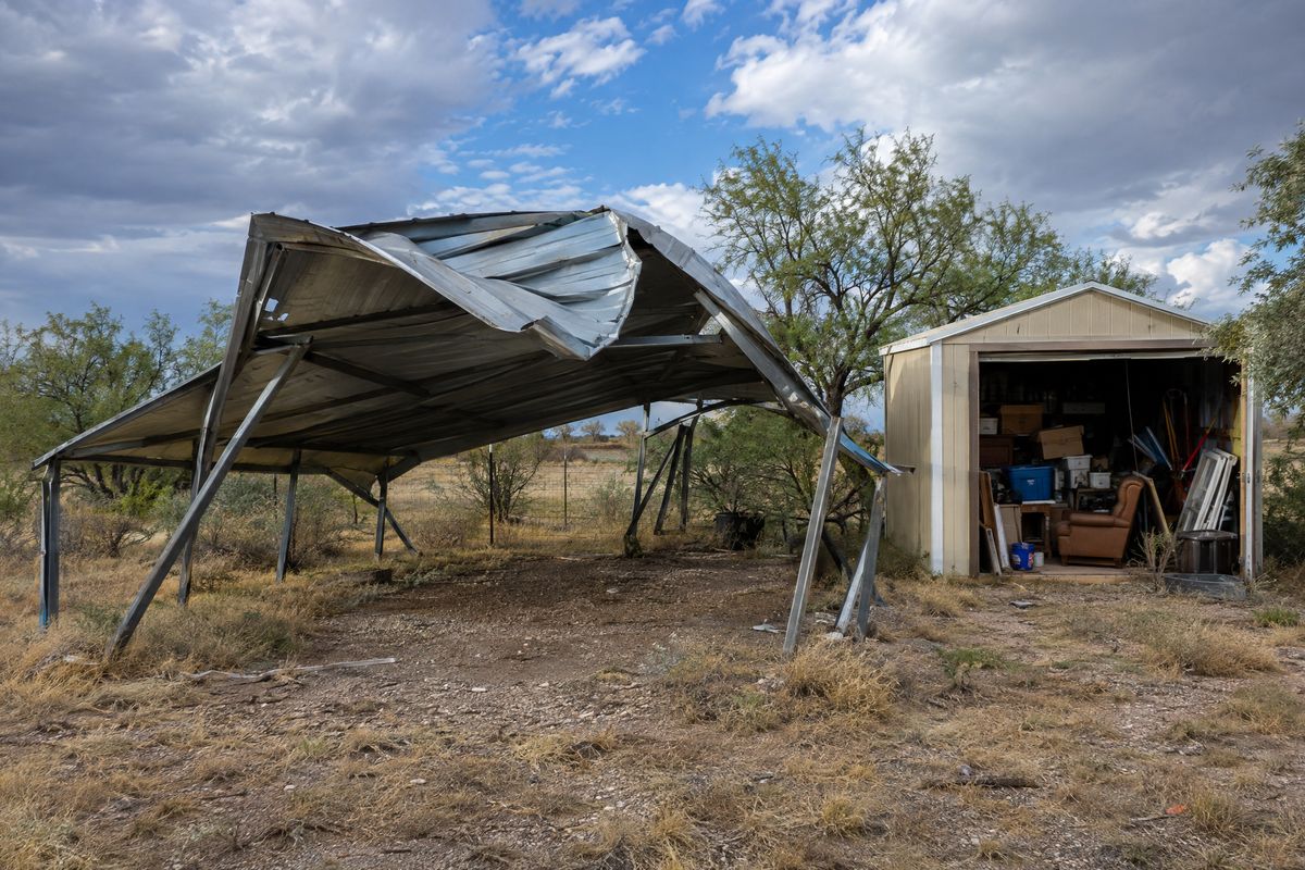 A wind-damaged metal carport with bent poles and a crumpled roof beside a storage shed overflowing with boxes and old furniture